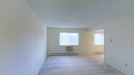 This single-floor space presents a clean and functional layout defined by its neutral color palette and practical features. The main living area is finished with wall-to-wall carpet flooring, offering a soft underfoot feel throughout. Crisp white walls and a smooth ceiling contribute to a bright, airy atmosphere. Natural light enters generously through multiple windows dressed with vertical blinds, allowing for adjustable light control while maintaining privacy. A wall-mounted air conditioning unit is positioned beneath one of the windows, complementing the baseboard heating system that runs along the perimeter of the room — together providing year-round climate comfort. An open doorway visible toward the far end of the space suggests additional rooms beyond, hinting at a functional floor plan. The overall ambiance is minimal and move-in ready, with a neutral foundation that lends itself well to a variety of interior styles. The space is well-lit, uncluttered, and benefits from a straightforward design that prioritizes utility and comfort in equal measure.