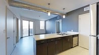 This contemporary single-floor unit showcases a thoughtfully designed open-concept layout that seamlessly connects the kitchen and living areas. The kitchen is a standout feature, anchored by a large central island with a light-toned countertop, double stainless steel sink, and sleek dark cabinetry offering ample storage. Pendant lighting hangs above the island, providing focused task illumination while adding a modern aesthetic touch. A stainless steel dishwasher completes the functional kitchen setup. Overhead, exposed silver spiral ductwork runs across the ceiling, lending an industrial character that contrasts stylishly with the cool blue-gray accent walls. The open living area flows naturally from the kitchen and is bathed in natural light courtesy of large sliding glass doors that lead out to a private balcony, offering outdoor views and a bright, airy atmosphere. The light-toned flooring throughout ties the space together with a clean, cohesive look. A doorway visible from the main living area connects to additional rooms, suggesting a well-organized and functional floor plan. The overall ambiance balances urban industrial design with modern residential comfort.