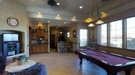 This single-floor space is designed with entertainment and comfort in mind, showcasing a seamless open layout that connects multiple functional areas. The game room serves as a central highlight, anchored by a full-size pool table beneath elegant pendant lighting that adds both style and practicality. Large windows allow natural light to fill the room, enhancing the warm, inviting ambiance throughout.

The wet bar area is a standout feature, complete with a raised counter, bar stool seating, and cabinetry, making it ideal for entertaining guests. Adjacent cabinetry with dark granite countertops provides additional storage and workspace. Arched doorways add an architectural charm reminiscent of Southwestern design, creating smooth transitions between spaces.

The flooring throughout is finished in large neutral ceramic tiles, providing a durable and cohesive look across the open floor plan. Recessed lighting is installed throughout the ceiling, complemented by a ceiling fan that ensures comfortable airflow. The warm, earthy tones of the walls and wood cabinetry contribute to a relaxed yet refined atmosphere. Multiple windows with blinds offer both natural illumination and privacy control, making this a well-appointed space suited for leisure and social gatherings alike.