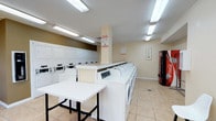 This shared laundry facility is designed for convenience and efficiency, offering a well-organized layout that accommodates multiple users simultaneously. The room features a generous row of front-loading washing machines aligned along one side, paired with a full bank of stacked dryers along the adjacent wall, providing ample capacity for residents. Bright fluorescent lighting installed across the ceiling illuminates the entire space evenly, creating a clean and functional environment. The light-toned walls and beige tile flooring contribute to the room's open and tidy appearance. A centrally placed folding table offers a dedicated area for sorting and folding laundry. Instructional and informational signage is posted throughout, including detergent usage guidelines, keeping users informed about proper machine operation. A vending machine near the entrance adds a layer of everyday convenience. The textured ceiling and practical layout reflect a straightforward, utilitarian design focused on serving the practical needs of its users. Overall, the space is well-maintained and efficiently arranged to support a smooth laundry experience.
