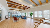 Located at 111 Teal Cir in Little River, South Carolina, this single-floor community space masterfully blends coastal charm with modern design. The open-plan layout seamlessly connects the kitchen and living areas, creating a welcoming, airy environment bathed in natural light from large windows and French doors that offer outdoor views.

Distinctive exposed wood ceiling beams in rich warm tones run the length of the space, adding architectural character against crisp white walls and a soft blue-accented ceiling. Light-toned hardwood flooring flows throughout, enhancing the bright, breezy atmosphere.

The kitchen is a standout feature, showcasing powder-blue beadboard cabinetry with brushed gold hardware, a generous white quartz island with bar seating, pendant lighting, and coastal-themed wall art. The open living area is thoughtfully appointed with a stylish seating arrangement, a statement chandelier, and lush tropical plants that reinforce the coastal aesthetic.

A dedicated dining nook with upholstered booth seating, wall sconces, and casual café-style tables provides an intimate spot for gathering. French doors connect the interior to outdoor areas, further expanding the sense of space. The overall ambiance is relaxed yet refined — a cohesive coastal retreat designed for community connection.