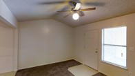 This single-floor space offers a straightforward and functional layout designed for comfortable everyday living. The main room is immediately welcoming, featuring vaulted ceilings that add a sense of height and openness to the interior. A classic five-blade ceiling fan with a built-in light fixture is centrally mounted, providing both air circulation and ambient lighting throughout the space. The room is finished with wall-to-wall carpeting in a neutral tone, offering a soft and cozy underfoot feel that complements the light-colored walls. A paneled entry door is positioned near a window fitted with blinds, allowing natural light to filter into the space while maintaining privacy. The textured ceiling adds a traditional architectural touch consistent throughout the room. The clean, neutral palette of the walls and flooring creates a versatile backdrop, making the space easy to personalize. Overall, the interior presents a practical and move-in-ready environment with classic residential finishes and a calm, understated ambiance suited to a variety of uses.