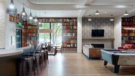 Located at 2981 Olentangy River Road in Columbus, Ohio, this single-floor community lounge is a thoughtfully designed gathering space with a distinct Ohio State University-inspired aesthetic. The open-concept layout accommodates a variety of social and recreational activities, seamlessly blending multiple functional zones within one generously sized room.

The space is immediately distinguished by its floor-to-ceiling bookshelf murals that line the walls, creating a warm, library-like atmosphere. Warm hardwood-style flooring runs throughout, grounding the industrial-chic design that incorporates exposed ductwork, white-painted brick columns, and textured stone accent walls.

A central bar counter with pendant lighting and metal bar stools serves as a natural gathering hub, complemented by a wall-mounted television set against a stacked stone backsplash. A cozy lounge area features a sleek linear fireplace framed by a textured tile surround and wood mantel, with comfortable seating including leather and upholstered chairs arranged around a metallic drum coffee table.

Large floor-to-ceiling windows flood the space with natural light and offer views of lush greenery outside. Additional entertainment options include a pool table and foosball table, making this a dynamic, multi-use destination for socializing and recreation. Multiple wall-mounted televisions ensure viewing access from nearly every corner of the space.