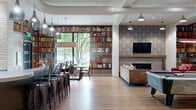Located at 2981 Olentangy River Road in Columbus, Ohio, this single-floor community lounge is a thoughtfully designed gathering space with a distinct Ohio State University-inspired aesthetic. The open-concept layout accommodates a variety of social and recreational activities, seamlessly blending multiple functional zones within one generously sized room.

The space is immediately distinguished by its floor-to-ceiling bookshelf murals that line the walls, creating a warm, library-like atmosphere. Warm hardwood-style flooring runs throughout, grounding the industrial-chic design that incorporates exposed ductwork, white-painted brick columns, and textured stone accent walls.

A central bar counter with pendant lighting and metal bar stools serves as a natural gathering hub, complemented by a wall-mounted television set against a stacked stone backsplash. A cozy lounge area features a sleek linear fireplace framed by a textured tile surround and wood mantel, with comfortable seating including leather and upholstered chairs arranged around a metallic drum coffee table.

Large floor-to-ceiling windows flood the space with natural light and offer views of lush greenery outside. Additional entertainment options include a pool table and foosball table, making this a dynamic, multi-use destination for socializing and recreation. Multiple wall-mounted televisions ensure viewing access from nearly every corner of the space.