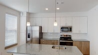 This single-story residence in Leander, Texas presents a clean, modern aesthetic throughout its well-designed layout. The open-concept kitchen serves as a central highlight, showcasing a large granite-topped island with an undermount sink and pendant lighting overhead. Cabinetry combines crisp white upper cabinets with warm wood-toned lower cabinets, complemented by a white subway tile backsplash. Stainless steel appliances, including a French door refrigerator, electric range, and over-the-range microwave, complete the kitchen's contemporary look. Light wood-style flooring extends through the main living areas, contributing to a bright and airy atmosphere, with natural light entering through windows fitted with white blinds and a glass-panel entry door.

The hallway features gray double panel doors providing access to additional spaces, including a closet for storage. Bedrooms are finished with plush carpet and ceiling fans, offering a comfortable, quiet retreat. One bedroom connects directly to a bathroom. The primary bathroom is elongated and well-appointed, featuring a dual-sink granite vanity with wood cabinetry, a large mirror with overhead lighting, tile flooring, and a separate shower area.