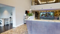 This elegant community amenity space is designed to impress from the moment you arrive. The grand lobby welcomes guests with a sleek reception desk topped with a marble-style surface, vibrant tropical florals, and herringbone-patterned hardwood flooring that flows throughout the main areas. Soaring double-height ceilings draw the eye upward to a mezzanine level adorned with geometric wrought-iron railings, adding architectural drama to the space. A large decorative aquarium serves as a striking focal point near the entrance corridor.

The expansive lounge area features oversized sectional seating in soft neutral tones, accented with rich blue throw pillows and a patterned area rug. A dramatic oversized clock anchors the wall above a bank of tall windows dressed with layered Roman shades and floor-length curtains, filling the room with natural light and outdoor views.

The full-service kitchen is outfitted with dark espresso cabinetry, stainless steel appliances, a teal herringbone tile backsplash, and marble countertops. A large communal dining table with bar-height seating accommodates gatherings of all sizes. An adjacent casual dining nook features built-in banquette seating with colorful tile detailing and compact café tables. Throughout the space, slate tile and herringbone wood flooring transitions define each zone while maintaining a polished, unified design.