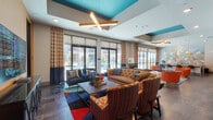 This dynamic single-floor community space is defined by its bold, contemporary interior design, combining a rich palette of teal, orange, and navy throughout. The main lounge area features floor-to-ceiling windows dressed with patterned curtains, flooding the space with natural light and providing views of the outdoor pool area beyond. Elevated tray ceilings with teal accents and distinctive cross-shaped pendant light fixtures add architectural interest overhead, while large-format tile flooring grounds the space throughout.

Multiple distinct seating zones create versatile gathering areas. A central lounge showcases a tufted caramel leather sofa paired with vibrant patterned accent chairs arranged around a wood coffee table atop a colorful area rug. A secondary seating alcove features deep navy wingback chairs flanking a rustic wood table, anchored by a wall-mounted flat-screen TV set against a textured wallcovering. A striking large-scale mosaic mural adds an artistic focal point near the entry corridor.

The game room area includes a billiards table, a round dining table with upholstered chairs, and an additional flat-screen TV mounted against a mixed-material mosaic tile accent wall. Throughout, curated artwork and layered lighting contribute to a lively yet sophisticated communal atmosphere.