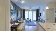 This single-floor residence offers an open-concept layout that seamlessly connects the dining and living areas, creating a cohesive and functional living environment. The living room is anchored by hardwood-style flooring and features a ceiling fan with a light fixture, complemented by track lighting that highlights the space. Large windows dressed with floor-length dark drapes allow generous natural light to filter in, enhancing the airy feel of the room. A sliding or French-style door provides access to an outdoor area, adding to the sense of openness.

The living area is thoughtfully arranged with a plush sofa, accent chair in teal, and a pair of round chrome-framed coffee tables atop a patterned area rug. A wall-mounted flat-screen television sits across from the seating arrangement. Bold abstract artwork adorns one wall, adding a striking visual focal point.

Adjacent to the living space, the dining area features a dark wood table with upholstered seating and elegant table settings. The nearby kitchen is partially visible, showcasing granite countertops. The overall ambiance is contemporary and welcoming, with a cool, neutral color palette throughout.