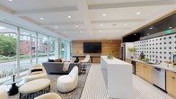 The Grand Flats Clubhouse is a thoughtfully designed single-floor amenity space that seamlessly blends contemporary style with functional comfort. The lobby greets visitors with polished concrete floors, a bold geometric tile pattern, and a striking gray accent wall bearing the Grand Flats branding. Resident mailboxes and elevator access are neatly integrated into this entry zone alongside a staffed management desk.

The main lounge and entertainment area is an open, light-filled space anchored by floor-to-ceiling windows that offer sweeping views of the surrounding streetscape. A coffered white ceiling, warm wood-slat accent wall, and a large wall-mounted flat-screen TV set the tone for relaxed gathering. A shuffleboard table adds a recreational focal point, while a variety of modern seating arrangements accommodate both casual socializing and quiet lounging.

The adjacent kitchen features sleek natural wood cabinetry, stone countertops, a stainless steel sink, under-counter refrigeration, and a bold cross-patterned tile backsplash. A secondary wet bar area with dark penny-tile accents provides additional beverage service capability.

Glass-walled conference and meeting rooms line a polished corridor, offering private spaces for work or small group gatherings. Warm wood flooring, curated wall art, and elegant pendant lighting complete these refined spaces. Throughout, the clubhouse balances an inviting residential warmth with a clean, modern aesthetic.