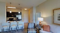 This single-floor residence offers a thoughtfully designed open-concept layout that seamlessly connects the kitchen and living areas, creating a welcoming and functional living environment. The modern kitchen is outfitted with dark cabinetry, stainless steel appliances including a French-door refrigerator, and a light-colored breakfast bar with seating — ideal for casual dining or entertaining. Pendant lighting above the bar adds a stylish touch, complementing the overall warm, neutral color palette found throughout the space. The living area is generously sized, featuring plush carpeting and room for comfortable furniture arrangements. A decorative accent chair with a colorful woven pattern, a sofa with warm-toned throw pillows, and a classic side lamp contribute to an inviting and relaxed ambiance. Smooth, light-toned walls and standard-height ceilings give the space a clean and airy feel. A hallway leads to additional rooms, with white-paneled interior doors providing a crisp, traditional aesthetic. The layout flows naturally from the entry through the main living spaces, making efficient use of the available footprint while maintaining a sense of openness and comfort throughout.