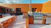 This rooftop terrace is a well-appointed outdoor amenity space designed for entertaining and relaxation, set atop a multi-story building with commanding panoramic views of the surrounding cityscape and horizon. The terrace is defined by bold, warm-toned orange cladding panels that line the perimeter walls, lending the space a vibrant, modern aesthetic that is both striking and cohesive.

At the heart of the terrace is a fully equipped outdoor kitchen, anchored by a stainless steel built-in grill and complementary appliances, all set beneath a decorative laser-cut pergola draped with bistro string lights. Dark granite countertops run the length of the kitchen island, providing ample prep and serving space. Orange powder-coated metal dining chairs and tables are positioned throughout the terrace, offering multiple seating areas for both casual and social gatherings.

A curved lounge area with a wall-mounted outdoor television creates an additional entertainment zone, ideal for relaxed gathering. Large-format porcelain floor tiles extend across the entire terrace, maintaining a clean, polished look underfoot. The open-air perimeter is lined with a secure metal railing, allowing unobstructed, sweeping views of the urban landscape stretching to the horizon — best appreciated during golden hour, when natural light bathes the terrace in warm tones.