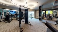This fitness center is a thoughtfully designed facility that combines modern aesthetics with a comprehensive selection of workout equipment. The space is defined by its distinctive interior design, featuring bold orange and blue vertical stripes on accent walls that create an energetic atmosphere, complemented by neutral gray tones throughout. Patterned carpet flooring runs across the entire facility, adding texture and visual interest underfoot.

The main workout area is illuminated by elegant circular pendant lighting fixtures and recessed ceiling lights, creating a well-lit environment. Large windows allow natural light to filter in, enhancing the open feel of the space. Full-length mirrors are strategically placed along the walls, visually expanding the room while allowing users to monitor their form.

The facility is comprehensively equipped with cardio machines including treadmills, stationary bikes, and ellipticals. A cable resistance machine serves as a centerpiece for strength training, accompanied by a dedicated free weights area featuring a multi-tiered dumbbell rack, a weight bench, and a barbell station. Functional training tools include plyometric boxes in various heights, stability balls, and adjustable dumbbells. Multiple wall-mounted flat-screen TVs are positioned throughout. A separate accessible restroom is located at the entrance alongside a keypad-secured glass door entry.