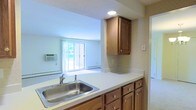 This single-floor residence offers a functional and straightforward layout that connects the kitchen and living area in an open, cohesive design. The kitchen is equipped with warm-toned wood cabinetry, both upper and lower, providing generous storage space. A double-basin stainless steel sink is set into a clean white countertop that extends along the kitchen peninsula, creating a natural division between the kitchen and the adjoining living space. Subtle patterned wallpaper adds a touch of character to the kitchen walls, complementing the classic cabinetry hardware. A recessed ceiling light provides focused illumination over the workspace. The living area beyond benefits from abundant natural light, courtesy of a large sliding glass door that opens to an outdoor view of lush greenery. A wall-mounted air conditioning unit and baseboard heating ensure year-round comfort. The space features textured ceilings throughout and is finished in soft, neutral tones that create a calm and inviting atmosphere. The overall layout is practical and well-suited for everyday living, with a seamless flow between the kitchen and the main living area.