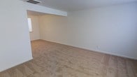 This two-floor property presents a straightforward and functional layout with a clean, neutral aesthetic throughout. The pictured main living area features wall-to-wall beige carpeting that extends across a generously sized open floor plan, creating a warm and understated foundation suitable for a variety of furnishing arrangements. The walls are finished in crisp white, contributing to a bright and airy atmosphere. A structural soffit detail is visible near the ceiling, adding subtle architectural dimension to the space. A ceiling-mounted HVAC vent ensures climate control is integrated directly into the room. Natural light is introduced through a window visible along one wall, supplementing the overall brightness of the interior. Neutral-toned baseboards line the perimeter, providing clean transitions between the walls and flooring. An electrical outlet is conveniently positioned along the far wall. The open, unobstructed layout of this room allows for flexible use, whether as a living room, recreation area, or multipurpose space. With its simple, move-in-ready condition and two-floor configuration, this property offers practical living across multiple levels.