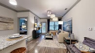 This thoughtfully designed single-floor residence offers a cohesive blend of modern finishes and comfortable living spaces throughout. The open-concept living room serves as the heart of the home, featuring wood-look plank flooring, crown molding, and large windows dressed with navy curtains that allow ample natural light. A stylish chandelier and arc floor lamp provide layered lighting, complementing the neutral color palette accented with cool blue tones.

The kitchen is equipped with dark espresso cabinetry, stainless steel appliances, a tile backsplash, and a granite-topped island with bar seating — ideal for casual dining or entertaining. Adjacent to the kitchen, a dedicated dining nook features a built-in display shelf and ample space for a full dining set.

The primary bedroom offers plush carpeting, a soothing teal accent wall, and access to an en-suite bathroom complete with a walk-in shower, a dark-framed vanity mirror, and a walk-in closet. The second bedroom echoes a similarly serene aesthetic with soft carpeting and a muted green accent wall. A second full bathroom includes a tub-shower combination with subway tile surround and wood-look flooring, maintaining a consistent design language throughout the residence.