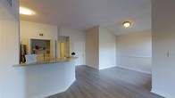 This single-floor space showcases an open-concept layout that seamlessly connects the kitchen and living areas. The kitchen features a curved granite-topped peninsula that serves as a natural divider, offering a practical breakfast bar while maintaining visual openness to the main living space. Light-toned cabinetry lines the kitchen walls, providing ample storage, and a pass-through window further enhances the open feel. The living area benefits from vaulted ceilings that create a heightened sense of space and airiness. Smooth, light grey wood-look flooring runs throughout, lending a clean and contemporary aesthetic that ties the spaces together cohesively. Neutral white walls keep the interior bright and versatile. A flush-mount ceiling light provides ambient illumination, while the overall layout is practical and easy to navigate. A white entry door is visible from the living area, offering a clear sense of the unit's flow. The combination of vaulted ceilings, open-plan design, and modern finishes creates a welcoming and functional environment suitable for everyday living.