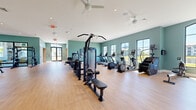 Located on Roma Way in Port Saint Lucie, Florida, this single-story fitness facility is designed to support a comprehensive wellness experience. The space is unified by a consistent design palette of soft teal-green walls, white ceilings, and light hardwood-style flooring, creating an energizing yet welcoming atmosphere throughout.

The main workout area is generously proportioned, accommodating a wide range of exercise equipment including multiple treadmills, elliptical machines, stair climbers, cable machines, selectorized strength equipment, free weights, benches, and a dedicated functional training rig. Large windows with black frames line the walls, flooding the interior with natural light and offering views of the surrounding community. Ceiling fans and recessed lighting maintain a comfortable environment.

A separate free weights and strength zone features a mirrored wall, a central dumbbell rack, and additional benches, providing an open layout suited for varied training routines. An adjoining studio room houses stability balls, kettlebells, resistance bands, and yoga mats for flexibility and functional fitness work.

The facility also includes a bright children's play area outfitted with a child-sized table and chairs, built-in shelving, a cushioned bench, and colorful décor, making the space family-friendly for community members.