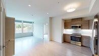 This contemporary single-floor residence showcases a clean, modern design characterized by crisp white walls, smooth light-toned hardwood flooring, and an open, airy layout throughout. The open-concept living and dining area is generously proportioned, bathed in natural light streaming through large windows that frame views of surrounding greenery, creating a bright and welcoming atmosphere.

The kitchen is thoughtfully designed with warm wood-grain flat-front cabinetry extending to the ceiling for ample storage, complemented by white countertops and a classic tile backsplash. A full suite of stainless steel appliances — including a range, over-the-range microwave, dishwasher, and french door refrigerator — provides a sleek, functional workspace well-suited for everyday living.

The bedroom is a spacious, light-filled retreat featuring wide windows with roll-down shades that overlook a lush green landscape, offering both privacy and a connection to the outdoors. The flooring continues seamlessly from the main living area, maintaining a cohesive aesthetic.

Two bathrooms serve the residence. A full bath includes a frameless glass-enclosed walk-in shower with clean white tile surround and brushed hardware. A secondary bathroom features a floating wood-toned vanity with a white countertop, a large mirror, and warm brushed gold accents. The overall palette is neutral and refined, lending the space a calm, modern ambiance throughout.