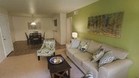 This single-floor residence offers a thoughtfully designed layout that blends comfort and functionality. The living area serves as the heart of the home, anchored by a striking green accent wall that adds a vibrant pop of color and warmth to the space. A plush sofa and patterned accent chair provide ample seating, while a dark wood coffee table grounds the room with a touch of contrast. Decorative throw pillows and wall art complement the green palette, creating a cohesive and welcoming ambiance. The living area flows naturally into a dedicated dining space, where a pendant light fixture hangs over a compact dining table and chairs, making the area ideal for shared meals. Hardwood-style flooring in the dining zone transitions into soft carpeting in the living area, adding texture and comfort underfoot. A hallway visible from the main living space leads to additional areas of the home, suggesting a practical and well-organized floor plan. The kitchen, partially visible, features light cabinetry and counter space suited for everyday cooking. Throughout the space, the interplay of natural tones, bold accents, and purposeful furnishings creates an atmosphere that feels both livable and stylishly curated.