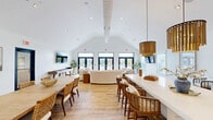 Located at 589 West Dr in Georgetown, Delaware, this single-floor community clubhouse offers a welcoming and versatile gathering space defined by its striking vaulted ceilings and crisp all-white interior. The open-plan layout is bathed in abundant natural light, streaming through multiple sets of large black-framed glass doors and transom windows that line the rear wall and provide direct views and access to an outdoor pool and recreational area.

The space is thoughtfully divided into distinct functional zones. A lounge area features plush, curved white sofas, wall-mounted flat-screen TVs, and a shuffleboard table, creating an inviting spot for relaxation and entertainment. Adjacent to the lounge, a long dining table with woven rattan chairs accommodates group gatherings beneath a pair of statement gold drum pendant lights that add warmth and elegance to the room.

The kitchenette area is well-appointed with light gray shaker-style cabinetry, dark granite countertops, a beverage refrigerator, and a generous white waterfall island that serves as both a workspace and a casual bar. Chalkboard-style coffee menus add a charming, café-inspired touch. Throughout the space, warm-toned luxury vinyl plank flooring ties every zone together, complemented by potted greenery that enhances the bright, airy ambiance of this well-designed community hub.