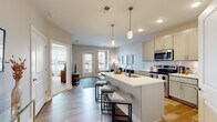 Located at 65 S. Washington Ave in Columbus, Ohio, this single-floor residence offers a thoughtfully designed open-concept layout with modern finishes throughout. The kitchen serves as a central hub of the living space, featuring gray shaker-style cabinetry, white quartz countertops, a full suite of stainless steel appliances, a subway tile backsplash, and a large island with bar seating illuminated by pendant lighting. Light wood-tone flooring flows through the kitchen and main living areas, contributing to a bright and cohesive aesthetic. The living room is generously sized, with multiple windows and a glass-paned door that invite abundant natural light and provide outdoor views. The space includes two well-proportioned bedrooms, each featuring large windows with blinds and plush carpeting. The primary bedroom connects directly to a private en-suite bathroom, which is finished with a single-sink vanity with quartz countertops, gray cabinetry, a framed mirror, and a walk-in glass-enclosed shower with white tile surround. Additional conveniences include an in-unit stacked washer and dryer. The overall design is clean and contemporary, with neutral tones creating a calm, welcoming atmosphere throughout.