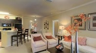 This single-floor residence offers a seamlessly connected living and dining area that exudes warmth and understated elegance. The living space is furnished with a pair of patterned accent chairs accented by terracotta throw pillows, complemented by a plush sofa and a dark wood coffee table that grounds the room. Decorative elements — including tall floral arrangements, framed wall art, and a mirror — add visual depth and a refined character to the space.

Crown molding runs along the ceiling, lending a polished architectural finish throughout. Warm ambient lighting from a combination of a stylish floor lamp and recessed ceiling fixtures creates an inviting, cozy atmosphere. The flooring transitions between carpet in the living area and what appears to be a harder surface near the dining space, offering both comfort and practicality.

The adjacent dining area is defined by a pendant light hanging above a dark dining table surrounded by matching chairs, creating a distinct yet open connection to the living room. A large mirror in the dining area amplifies the sense of space and reflects the warm tones carried throughout. Together, the rooms present a cohesive, well-appointed interior that balances comfort with a tasteful, contemporary aesthetic.