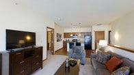 This single-floor residence features an open-concept layout that connects the living area and kitchen, creating a cohesive and functional living environment. The living space is finished with plush carpeting and is furnished with comfortable gray upholstered seating arranged around a dark wood coffee table, with a large flat-screen television positioned on a matching media console. Warm-toned wood baseboards complement the neutral white walls throughout.

The kitchen is positioned at the rear of the open space and features rich, dark wood cabinetry paired with black appliances, including a refrigerator, stove, and microwave. A peninsula-style counter serves as a natural divider between the kitchen and the living area, providing additional workspace and casual seating potential. Pendant lighting hangs above the peninsula, adding both function and a modern aesthetic touch.

The flooring transitions from carpet in the living area to hardwood-style flooring in the kitchen zone, clearly delineating the two spaces while maintaining visual continuity. A hallway visible from the main living area leads to additional rooms, suggesting a thoughtfully designed layout with clear separation between living and private spaces.