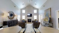 This single-story space is defined by its striking vaulted ceilings, which rise dramatically to a peak and are complemented by a ceiling fan and recessed lighting that illuminate the open interior. The great room serves as the focal point, anchored by a classic white-mantled fireplace with a tile surround, above which a flat-screen television is mounted. Multiple windows positioned at both standard and elevated heights flood the space with abundant natural light, enhancing the airy, open atmosphere created by the soaring ceiling height. The neutral, warm-toned walls provide a clean backdrop that complements the light hardwood flooring throughout. The living area is furnished with plush, tufted brown sofas and armchairs arranged symmetrically around a patterned area rug in soft blue and grey tones, encouraging conversation and relaxation. A dark wood credenza with decorative metalwork adds a refined accent along one wall. To the right, a secondary seating or display area with a glass-topped console provides additional functionality. Overall, the space conveys an elegant yet approachable ambiance suited to both everyday use and entertaining.