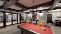 This billiards lounge is a well-appointed recreational space designed for both casual play and relaxed socializing. The centerpiece is a full-size pool table with a striking red felt surface, positioned beneath a dedicated billiard light fixture with multiple pendant shades that provide focused, even illumination over the playing surface. The ceiling throughout features dark wood-trimmed coffered beamwork, lending the space a refined, club-like character that is carried through in the rich wood baseboards and trim details.

Adjacent to the pool table, a comfortable seating area includes yellow upholstered armchairs arranged around a dark wood console table adorned with framed artwork and matching table lamps, creating an inviting lounge atmosphere. A small dining area with a round table and dark chairs offers additional gathering space nearby.

A built-in bar area with a granite-topped counter and dark leather barstools provides a social hub for refreshments. A wall-mounted television accompanied by a counter-height bar ledge and additional seating creates a dedicated viewing area. Neutral carpet flooring and light grey walls maintain a clean backdrop throughout, while wall sconces offer warm ambient lighting. A vestibule area with modern chaise lounge seating provides a transitional space near the entrance.