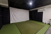 Located at 225 Sandwich St N in Amherstburg, Ontario, this purpose-built indoor golf simulator space offers a fully equipped and climate-controlled environment for year-round golf practice and entertainment. The single-floor space is thoughtfully designed to maximize functionality within a compact footprint.

At the heart of the room is a professional-grade artificial turf hitting area, featuring a raised hitting mat with multiple tee positions set into the green surface. A large, floor-to-ceiling white projection screen dominates the front wall, serving as the display surface for the golf simulator. The surrounding walls are lined with floor-to-ceiling padded black panels, providing both impact protection and acoustic dampening to create an immersive playing environment.

The space transitions seamlessly into a lounge and viewing area, where a rustic wooden communal table paired with industrial-style adjustable stools provides comfortable seating for guests. A ceiling-mounted projector is positioned overhead, and a wall-mounted mini-split air conditioning unit ensures consistent climate control throughout. A compact white cabinetry unit with a granite-style countertop offers convenient surface space. The dark tiled drop ceiling, recessed lighting, and grey carpet tiles in the lounge area round out the polished, functional aesthetic of this well-appointed simulator bay.
