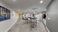 The Media Lounge at 150 Southfield Ave in Stamford, Connecticut is a thoughtfully designed single-floor amenity space that blends comfort, functionality, and modern aesthetics. The layout flows seamlessly from one zone to the next, offering a variety of seating configurations to suit different activities and group sizes.

At the heart of the lounge, a relaxed conversation area features sleek leather sofas arranged around a solid wood coffee table, anchored by a patterned area rug. Adjacent to this is a café-style dining section with multiple high-top round tables paired with contemporary white and chrome bar stools, ideal for casual gatherings or individual work sessions. Pendant lighting and recessed ceiling fixtures provide layered illumination throughout.

The space features a consistent palette of light gray large-format tile flooring, crisp white walls, and contrasting charcoal accent walls that add depth and visual interest. Colorful framed artwork is displayed throughout, lending a gallery-like quality to the corridors and open areas. A beverage station with countertop appliances serves as a practical amenity hub.

Natural light filters in through large sliding glass doors and windows along one end of the lounge, offering views of the surrounding greenery. Wide, well-lit hallways connect the various zones, ensuring easy navigation. The overall ambiance is clean, contemporary, and welcoming — well-suited for both social interaction and focused individual use.