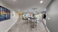 The Media Lounge at 150 Southfield Ave in Stamford, Connecticut is a thoughtfully designed single-floor amenity space that blends comfort, functionality, and modern aesthetics. The layout flows seamlessly from one zone to the next, offering a variety of seating configurations to suit different activities and group sizes.

At the heart of the lounge, a relaxed conversation area features sleek leather sofas arranged around a solid wood coffee table, anchored by a patterned area rug. Adjacent to this is a café-style dining section with multiple high-top round tables paired with contemporary white and chrome bar stools, ideal for casual gatherings or individual work sessions. Pendant lighting and recessed ceiling fixtures provide layered illumination throughout.

The space features a consistent palette of light gray large-format tile flooring, crisp white walls, and contrasting charcoal accent walls that add depth and visual interest. Colorful framed artwork is displayed throughout, lending a gallery-like quality to the corridors and open areas. A beverage station with countertop appliances serves as a practical amenity hub.

Natural light filters in through large sliding glass doors and windows along one end of the lounge, offering views of the surrounding greenery. Wide, well-lit hallways connect the various zones, ensuring easy navigation. The overall ambiance is clean, contemporary, and welcoming — well-suited for both social interaction and focused individual use.