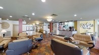 This single-floor lobby presents a well-organized and functional hospitality space designed to accommodate guests with comfort and convenience. The seating area features plush black and beige leather sofas and armchairs arranged around sleek glass-top coffee tables, providing a relaxed setting for guests to unwind. The flooring showcases a bold, patterned carpet with swirling motifs in rich tones of brown, gold, and red, adding a distinctive visual character to the space.

Recessed lighting and pendant fixtures illuminate the area evenly, complementing the light-colored ceiling and walls. A café or refreshment counter is positioned toward one side, equipped with coffee machines and service displays, offering guests easy access to beverages. Nearby, a front service desk with a tiled counter serves as the main reception area, accented by teal cabinetry above and warm lighting.

Further into the space, a secondary lounge area with bar-height seating and small bistro tables is visible, adding versatility to the layout. Natural light filters in from what appears to be a glass entrance in the background, enhancing the open and welcoming ambiance throughout the lobby.