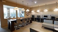 This single-floor amenity space combines casual social areas with functional workspaces in a warm, inviting environment. The open layout is anchored by a long bar-height communal table lined with sleek, upholstered swivel stools featuring polished chrome bases, illuminated by a row of elegant white pendant lights that hang at varying heights overhead. Large windows along one wall allow natural light to fill the seating area, complementing the warm beige tones throughout the space. A decorative metallic wall sculpture adds an artistic focal point between the seating and work zones. Along the opposite wall, a dedicated computer workstation area features built-in desks set against a textured stacked-stone accent wall, providing an organized and private setting for individual use. Each workstation is equipped with a desktop computer and an ergonomic task chair. Recessed ceiling lighting supplements the pendant fixtures, ensuring consistent illumination throughout. The neutral color palette of tan, beige, and brown, combined with the modern furnishings, creates a cohesive and comfortable atmosphere suited for both focused work and relaxed social interaction.
