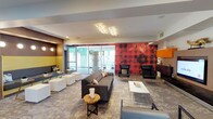 This single-floor lounge space makes a striking first impression with its bold, eclectic interior design. The open layout flows seamlessly between several distinct seating areas, creating a dynamic yet cohesive environment. A standout feature is the oversized red plaid floral accent wall, which anchors one end of the room and adds a dramatic pop of color alongside vibrant orange, yellow, and magenta wall-mounted cabinetry elements. A large flat-screen television is integrated into a warm wood-panel media wall, flanked by sculptural gold décor pieces, making it a natural focal point for gathering. Seating is plentiful and varied, ranging from a tufted gray sofa and sleek black leather lounge chairs to modular white cube ottomans and a cozy built-in banquette nook with a yellow backing, globe-style sconce lighting, and small potted plants. Patterned carpeting in muted gray and gold tones runs throughout, adding texture underfoot. Large frosted glass sliding doors allow natural light to filter in while maintaining privacy. A uniquely designed cluster ceiling fixture adds an artistic touch overhead. The space opens toward additional rooms visible in the background, suggesting further amenities beyond the main lounge area.