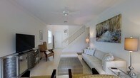 This two-story home offers a clean, coastal-inspired aesthetic defined by a predominantly white and neutral color palette throughout. The main living area serves as the heart of the ground floor, featuring light-toned tile flooring that extends across the space and contributes to an airy, open atmosphere. A ceiling fan overhead provides both function and a relaxed, casual touch to the room.

The living area is well-appointed with comfortable seating, including a plush white sofa accented with decorative pillows and complemented by natural wicker-style accent chairs — a nod to a relaxed, coastal sensibility. A textured metallic coffee table anchors the seating arrangement, while a large flat-screen television sits opposite. Wall art adds visual interest, with a prominent abstract painting in muted tones displayed prominently on one wall, and a framed piece near the entryway adding further character.

A white-painted staircase with open balusters rises gracefully from the main living space, connecting to the second floor and serving as a subtle architectural focal point. Natural light filters in through a window near the entry, brightening the space. The overall ambiance is calm, cohesive, and inviting, with a design language that balances simplicity and warmth across both levels of the home.