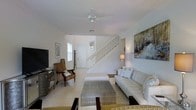 This two-story home offers a clean, coastal-inspired aesthetic defined by a predominantly white and neutral color palette throughout. The main living area serves as the heart of the ground floor, featuring light-toned tile flooring that extends across the space and contributes to an airy, open atmosphere. A ceiling fan overhead provides both function and a relaxed, casual touch to the room.

The living area is well-appointed with comfortable seating, including a plush white sofa accented with decorative pillows and complemented by natural wicker-style accent chairs — a nod to a relaxed, coastal sensibility. A textured metallic coffee table anchors the seating arrangement, while a large flat-screen television sits opposite. Wall art adds visual interest, with a prominent abstract painting in muted tones displayed prominently on one wall, and a framed piece near the entryway adding further character.

A white-painted staircase with open balusters rises gracefully from the main living space, connecting to the second floor and serving as a subtle architectural focal point. Natural light filters in through a window near the entry, brightening the space. The overall ambiance is calm, cohesive, and inviting, with a design language that balances simplicity and warmth across both levels of the home.