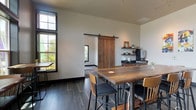 This single-story communal lounge blends modern industrial design with rustic warmth, creating an inviting gathering space suited for casual meetings and socializing. The interior is anchored by dark slate-style tile flooring, which contrasts handsomely with the reclaimed wood plank accent wall that runs along one side of the room, adding texture and character throughout.

Seating is arranged around multiple high-top communal tables crafted from solid wood, paired with a mix of bar-height chairs in black and natural wood tones. Along one wall, tufted banquette seating in a muted teal-gray provides a comfortable lounge alternative. Track lighting mounted on the ceiling offers flexible, well-distributed illumination across the space.

Large black-framed windows line multiple walls, flooding the interior with natural light and offering views of the surrounding outdoor environment. A sliding barn door, constructed from warm-toned reclaimed wood and hung on black hardware, serves as a striking architectural detail while providing access to an adjacent area.

A dedicated coffee station features wood cabinetry with a dark countertop, open floating shelves, and a Starbucks-branded coffee setup. Colorful abstract artwork displayed on the white walls adds a curated, contemporary touch, completing the space's modern yet approachable aesthetic.