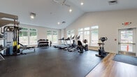 Located at 702 Pleasant Valley Rd N in Groton, Connecticut, this dedicated fitness center is a single-story facility designed to support a comprehensive workout experience. The main exercise area is highlighted by soaring vaulted ceilings with recessed lighting and a ceiling fan, creating an open, airy atmosphere. Large white-framed windows line the walls, flooding the space with natural light and offering views of the surrounding wooded landscape.

The workout floor features durable rubber flooring throughout the primary exercise zone, transitioning to hardwood near the entrance area. Equipment includes multiple treadmills, an elliptical machine, a stationary upright bike, a recumbent bike, and a multi-station cable machine with a functional training rack. A tiered dumbbell rack offers a full range of free weights for strength training. The layout provides ample open floor space for stretching and floor exercises.

Supporting amenities include restrooms with clearly marked entrances along the back wall, a water fountain, and a first aid station mounted near the exit. A glass-paneled exterior door provides direct access to the outdoors. The facility is equipped with a fire alarm system and clearly posted exit signage, ensuring safety throughout. The clean, neutral color palette and organized equipment arrangement contribute to a professional and functional environment.