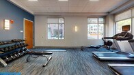 Located at 160 Pleasant St in Malden, Massachusetts, this well-equipped fitness center offers a comprehensive single-floor workout facility designed to support a wide range of exercise routines. The space features a clean, modern aesthetic with neutral walls accented by bold blue panels, a drop tile ceiling with recessed lighting, and durable patterned carpet flooring throughout.

Large windows line multiple walls, flooding the interior with natural light and providing views of the surrounding streetscape, creating an open and energizing atmosphere. The cardio zone is anchored by a row of treadmills equipped with individual screens, accompanied by elliptical machines and a rowing machine, all positioned to take advantage of the natural light from the windows. A wall-mounted flat-screen television serves the cardio area for added entertainment.

The strength and functional training section includes a dual cable machine, a weight scale, a rack of dumbbells in varying sizes, and a padded workout bench. A dedicated stretching and functional fitness area features stability balls in multiple sizes, resistance bands, medicine balls, and exercise mats, providing a versatile space for dynamic workouts. Wall sconces add warm accent lighting, complementing the bright overhead panels. The facility is thoughtfully organized to maximize usable floor space while keeping equipment accessible and well-arranged.