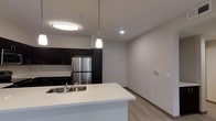 This single-floor space showcases a clean, modern design with an open-concept layout that seamlessly connects the kitchen and living areas. The kitchen is equipped with dark espresso-toned cabinetry that extends to the ceiling, providing ample storage, and is paired with stainless steel appliances, including a full-size refrigerator. A white quartz-style kitchen island anchors the space, featuring an undermount sink and offering additional workspace and casual seating potential. A contemporary pendant light hangs above the island, complementing the flush-mount ceiling fixture and recessed lighting that together provide layered illumination throughout the area. The flooring consists of light wood-look planks that run continuously through the kitchen and into the adjoining living space, adding warmth while maintaining a cohesive aesthetic. Neutral grey walls keep the interior feeling bright and versatile. A wall-mounted thermostat is visible in the living area, indicating modern climate control. The overall ambiance is clean, minimal, and move-in ready, with a practical layout suited for everyday living.