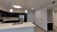 This single-floor space showcases a clean, modern design with an open-concept layout that seamlessly connects the kitchen and living areas. The kitchen is equipped with dark espresso-toned cabinetry that extends to the ceiling, providing ample storage, and is paired with stainless steel appliances, including a full-size refrigerator. A white quartz-style kitchen island anchors the space, featuring an undermount sink and offering additional workspace and casual seating potential. A contemporary pendant light hangs above the island, complementing the flush-mount ceiling fixture and recessed lighting that together provide layered illumination throughout the area. The flooring consists of light wood-look planks that run continuously through the kitchen and into the adjoining living space, adding warmth while maintaining a cohesive aesthetic. Neutral grey walls keep the interior feeling bright and versatile. A wall-mounted thermostat is visible in the living area, indicating modern climate control. The overall ambiance is clean, minimal, and move-in ready, with a practical layout suited for everyday living.