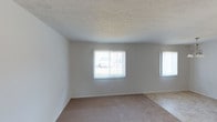 This single-floor space presents a clean, functional layout with a straightforward design. The main living area is finished with wall-to-wall beige carpeting that extends throughout, providing a neutral and comfortable foundation underfoot. The walls are painted in a light, neutral tone, creating a bright and open atmosphere that complements the natural light flowing in through multiple windows. Two windows fitted with vertical blinds line one wall, allowing for adjustable light control while offering views to the exterior. The textured ceiling adds a subtle visual detail to the otherwise understated interior. The room transitions toward what appears to be a tiled area, suggesting a defined boundary between the living space and an adjoining functional zone such as a dining or kitchen area. Overall, the space maintains a simple, move-in-ready aesthetic with neutral finishes throughout, offering a versatile canvas that can be easily personalized. The abundance of natural light and light color palette contribute to an airy, open feel throughout the interior.