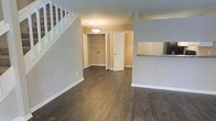 This two-story space is designed with a clean, modern aesthetic that balances functionality and comfort. The main level showcases rich, dark hardwood-style flooring that runs throughout, providing a striking contrast against the soft gray walls and crisp white trim and baseboards. The open layout of the main living area allows for easy flow between spaces, creating a sense of openness and versatility.

A staircase with white balusters and railings is prominently positioned, adding a classic architectural element while providing access to the upper level. A pendant light fixture hangs from the ceiling, offering focused ambient lighting over the central area. Adjacent to the living space, a bifold-door closet provides convenient built-in storage, while a separate door leads to an additional room beyond.

To one side, a partial view of the kitchen reveals a raised countertop with granite-style surfaces and a stainless steel refrigerator, hinting at a well-appointed culinary space. The overall ambiance is neutral, clean, and move-in ready, with a cohesive color palette and thoughtful design details throughout the main level.