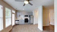 This well-appointed studio offers a thoughtfully designed open-concept layout that maximizes every square foot. The kitchen serves as a central highlight, featuring sleek gray cabinetry, a classic white subway tile backsplash, and light-toned countertops that create a clean, contemporary aesthetic. A full suite of stainless steel appliances — including a refrigerator, electric range, built-in microwave, and dishwasher — provides all the essentials for comfortable day-to-day living. Light wood-style plank flooring runs throughout the main living area, lending warmth and continuity to the space. A ceiling fan overhead ensures efficient air circulation year-round, complementing the recessed lighting that keeps the interior bright and inviting. Natural light filters in through the windows, enhancing the open and airy atmosphere. A clean white column and partial wall subtly define the transition between the kitchen and living areas while preserving the open feel. A private door leads to an additional area, offering functional separation within the layout. The overall design is modern and move-in ready, balancing style with practicality.