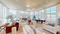 Located at 200 Big Woods Drive in Poquoson, Virginia, this single-floor community space offers a generously sized, open-concept layout designed for gathering and relaxation. The interior is defined by soaring ceilings and floor-to-ceiling windows that wrap around the perimeter, flooding the space with abundant natural light and providing sweeping views of the surrounding neighborhood.

The main area is thoughtfully organized into distinct functional zones. A comfortable lounge area features multiple seating arrangements anchored by a round wood coffee table and striped ottomans, centered beneath a pair of oversized pendant lights. Adjacent to this is a formal dining area with a large rectangular table and modern chairs, ideal for group gatherings. A built-in wet bar with white cabinetry, a wood-accented floating shelf, and a decorative tile backsplash provides a convenient refreshment station.

Elegant arched doorways frame transitions between areas, lending a classic architectural character to the otherwise contemporary design. Light-toned large-format tile flooring runs throughout, enhancing the airy, cohesive aesthetic. A dedicated nook near the entry features curated artwork, wall sconces, and a media console, adding a welcoming and refined touch. The overall ambiance is bright, clean, and inviting—a polished gathering space that balances comfort with sophisticated design.