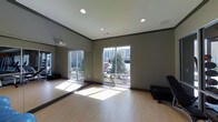 This yoga room is thoughtfully designed to support focused practice and movement. The space features smooth hardwood-style flooring that provides a clean, comfortable surface ideal for yoga and stretching exercises. Large sliding windows along one wall flood the room with abundant natural light, offering views of surrounding greenery and the exterior landscape, creating a calming, connected atmosphere. Full-length mirrors line an adjacent wall, allowing practitioners to monitor alignment and form during sessions. Recessed ceiling lights provide even, adjustable illumination for use during early morning or evening classes when natural light is limited. The ceiling height adds a sense of openness and airiness to the room, preventing any feeling of confinement during practice. Neutral-toned walls in a soft gray palette contribute to a serene and distraction-free environment. The open floor plan maximizes usable space, accommodating both individual sessions and small group classes. Overall, the room strikes a balance between functional design and a tranquil ambiance well-suited for yoga, meditation, and wellness activities.