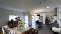 This single-floor space showcases a sleek, modern design built around an open-concept layout that connects the living and kitchen areas in one cohesive, flowing environment. The living area features rich dark hardwood flooring, smooth light-gray walls, and recessed lighting that complements the natural light streaming in through large sliding glass doors. These doors open to what appears to be an outdoor balcony or deck, further enhancing the sense of openness and space. The living area is appointed with a leather sofa, a glass-top coffee table, and curated décor pieces, creating a comfortable yet refined atmosphere. Transitioning seamlessly into the kitchen, the space is anchored by a central island with granite countertops and black bar seating, making it ideal for casual dining or entertaining. The kitchen features crisp white cabinetry extending to the ceiling, a stainless steel refrigerator, built-in microwave, and a bold black pendant light that adds a dramatic focal point overhead. Throughout the space, the contrast of dark flooring against white cabinetry and light walls creates a striking visual balance. The open sightlines, modern finishes, and thoughtful layout combine to deliver a space that is both highly functional and visually sophisticated.