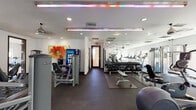 This fitness center is thoughtfully organized into two primary zones, offering a comprehensive workout experience. The cardio and strength training area features rubber flooring and is outfitted with a full row of treadmills, elliptical machines, and cable machines, complemented by a complete dumbbell rack with weight bench and a selection of strength training equipment. Floor-to-ceiling mirrors line one wall, enhancing the sense of space throughout this section. Recessed lighting and colorful LED accent strips illuminate the area, while ceiling fans ensure comfortable airflow. A beverage vending machine provides added convenience.

The adjacent studio area features warm hardwood flooring and is bathed in natural light through a series of large windows that frame outdoor views of mature trees and the surrounding landscape. This open space accommodates spinning bikes, medicine balls, stability balls, and a yoga mat area, making it ideal for group or individual training sessions. A wall-mounted flat-screen TV and a glass partition wall connecting to the main gym area complete the studio. The clean, white walls and modern finishes throughout give the fitness center a bright, professional ambiance suited for a wide range of fitness activities.