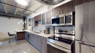 This single-floor loft masterfully blends industrial character with contemporary design. Exposed silver ductwork and structural elements run throughout the ceiling, creating an authentic urban aesthetic that contrasts beautifully with the clean, modern finishes below. Polished terrazzo floors flow through the hallways and primary living areas, adding texture and a reflective quality to the space.

The kitchen is a standout feature, outfitted with dark wood-grain cabinetry, frosted glass upper cabinets, a textured mosaic tile backsplash, quartz countertops, and a full suite of stainless steel appliances including a range, microwave, and dishwasher. A large island with seating for multiple guests bridges the kitchen and dining area, illuminated by striking pendant cluster lighting that adds a dramatic focal point overhead.

The living area offers a relaxed, cozy atmosphere with neutral tones and exposed ceiling infrastructure maintaining the loft's industrial identity. The bedroom is a calm retreat, featuring a closet and the same exposed ceiling details that carry throughout the unit.

The hallway connects rooms with polished floors, white-paneled doors, and a sliding barn door accent. The bathroom is particularly noteworthy, showcasing exposed original brick walls, a large window with natural light, a backlit vanity mirror, quartz countertop, and polished terrazzo flooring — a striking fusion of raw and refined materials.